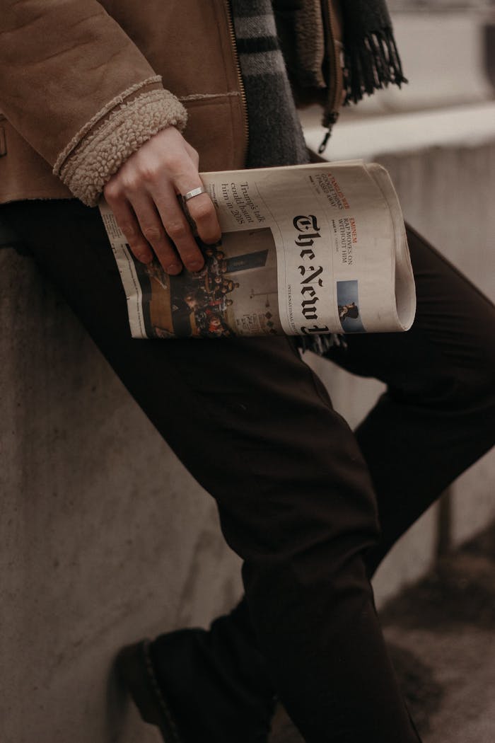 A fashionable man holds a newspaper while leaning against a city wall, showcasing modern urban style.